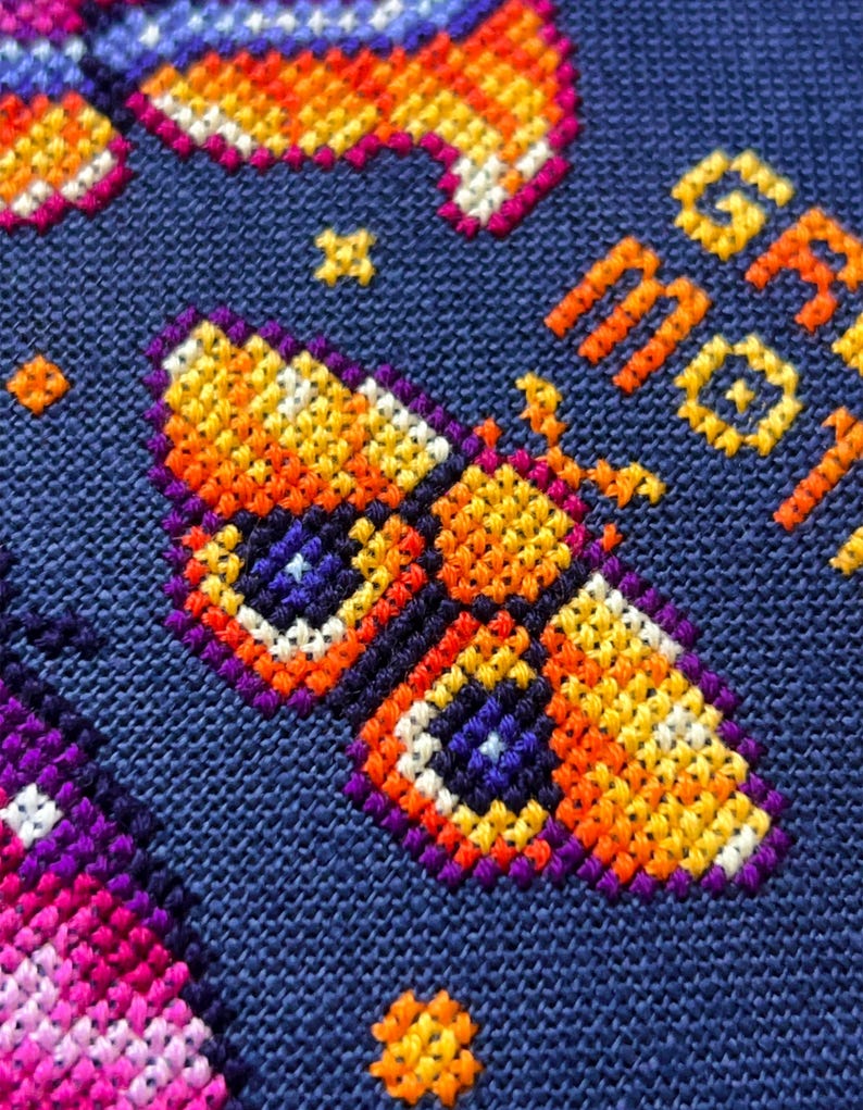 Op de afbeelding: Close-up van een kruissteekontwerp met een kleurrijke vlinder met oranje, gele en paarse tinten op een donkerblauwe achtergrond. De tekst "GR MOT" is in oranje gestikt. Het ontwerp bevat sterren en andere geometrische vormen.