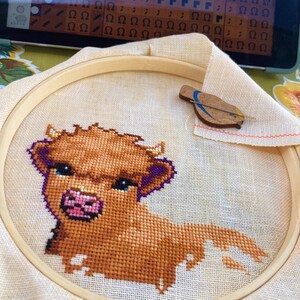 May include: A close-up of a brown and white cross-stitch embroidery of a Highland cow with purple accents. The embroidery is in a wooden embroidery hoop.