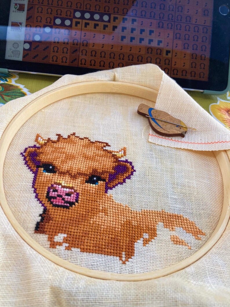 May include: A close-up of a brown and white cross-stitch embroidery of a Highland cow with purple accents. The embroidery is in a wooden embroidery hoop.