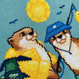 May include: Cross-stitch artwork depicting two otters, one in a yellow raincoat and the other in a blue hat, fishing under a yellow sun. The background is a light blue fabric, with small blue birds. The otters are wearing glasses.