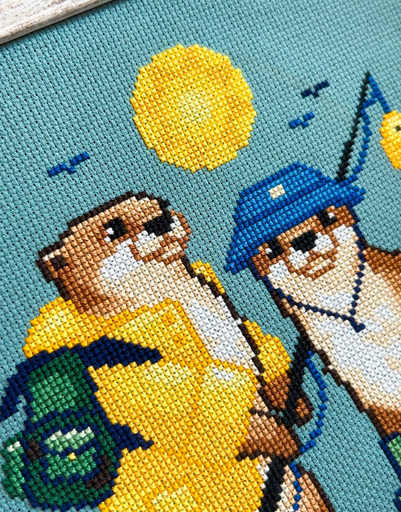 May include: Cross-stitch artwork depicting two otters, one in a yellow raincoat and the other in a blue hat, fishing under a yellow sun. The background is a light blue fabric, with small blue birds. The otters are wearing glasses.
