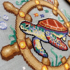 May include: A colorful cross-stitch design featuring a sea turtle with a brown shell and a blue, green, and yellow patterned body. The turtle is swimming in a circle of brown wood with yellow accents, surrounded by blue bubbles and green seaweed.