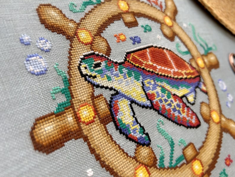 May include: A colorful cross-stitch design featuring a sea turtle with a brown shell and a blue, green, and yellow patterned body. The turtle is swimming in a circle of brown wood with yellow accents, surrounded by blue bubbles and green seaweed.