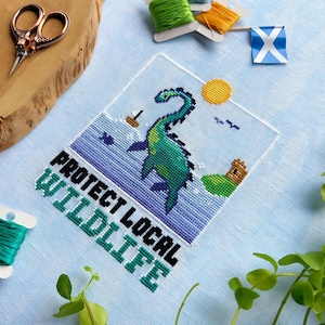 May include: Cross-stitch pattern featuring a green and blue Nessie, the Loch Ness Monster, with the text "Protect Local Wildlife".