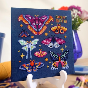 Op de afbeelding: Een kruissteekkunstwerk met diverse kleurrijke motontwerpen op een marineblauwe achtergrond. De zin "GREAT MOTHS!" is in oranje gestikt. Het kunstwerk wordt op een witte ezel gepresenteerd.