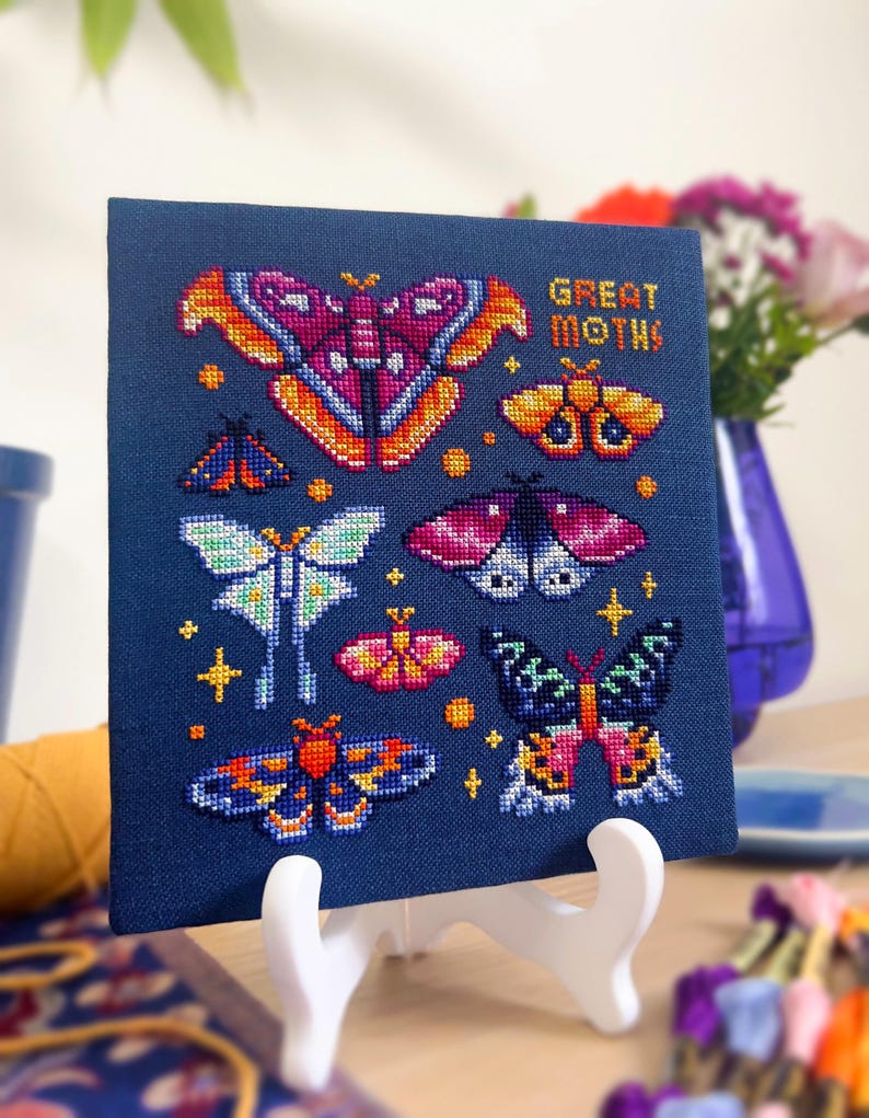 Op de afbeelding: Een kruissteekkunstwerk met diverse kleurrijke motontwerpen op een marineblauwe achtergrond. De zin "GREAT MOTHS!" is in oranje gestikt. Het kunstwerk wordt op een witte ezel gepresenteerd.