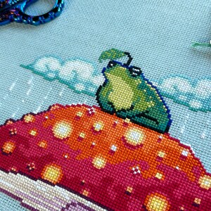 May include: A green frog with a leaf on its head sits on a red and orange mushroom with white spots. The background is a light blue sky with white clouds and rain.