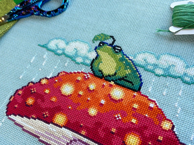 May include: A green frog with a leaf on its head sits on a red and orange mushroom with white spots. The background is a light blue sky with white clouds and rain.
