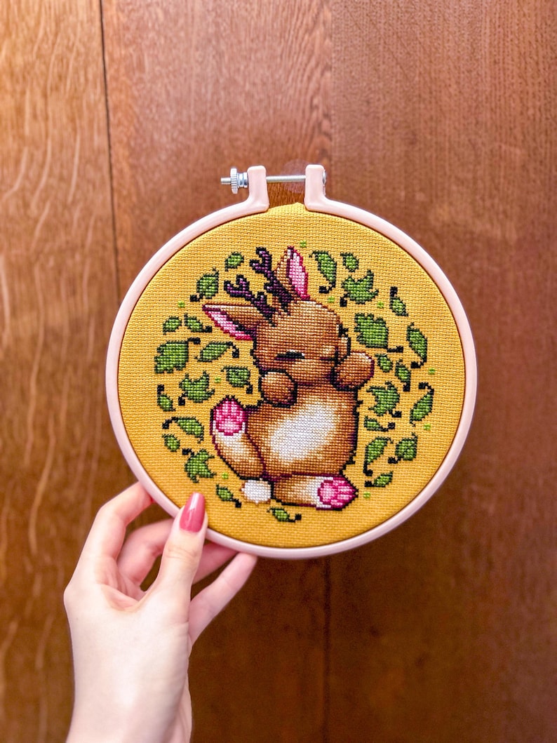 May include: A yellow embroidery hoop with a pink outer ring, featuring a cross-stitch design of a brown deer with pink accents, surrounded by green leaves.