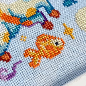 May include: Cross-stitch embroidery featuring a rocket ship and a goldfish against a light blue background. The rocket ship is detailed with orange, blue, and yellow threads. The goldfish is orange and yellow. Other elements include stars and clouds.