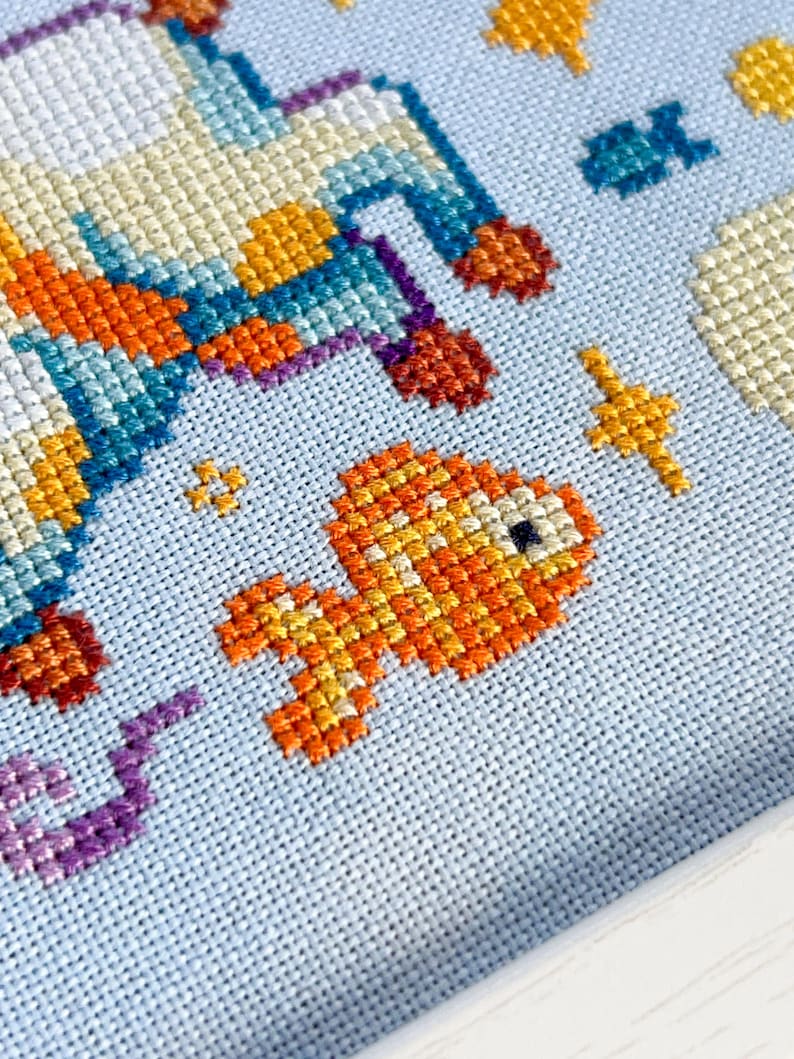 May include: Cross-stitch embroidery featuring a rocket ship and a goldfish against a light blue background. The rocket ship is detailed with orange, blue, and yellow threads. The goldfish is orange and yellow. Other elements include stars and clouds.