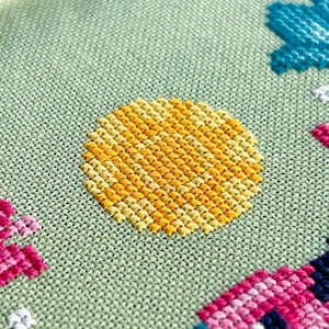 May include: Close-up of a cross-stitch design on a light green fabric. The design features a yellow sun, a blue flower, and a pink house. The stitches are neatly aligned, creating a textured and colorful pattern.