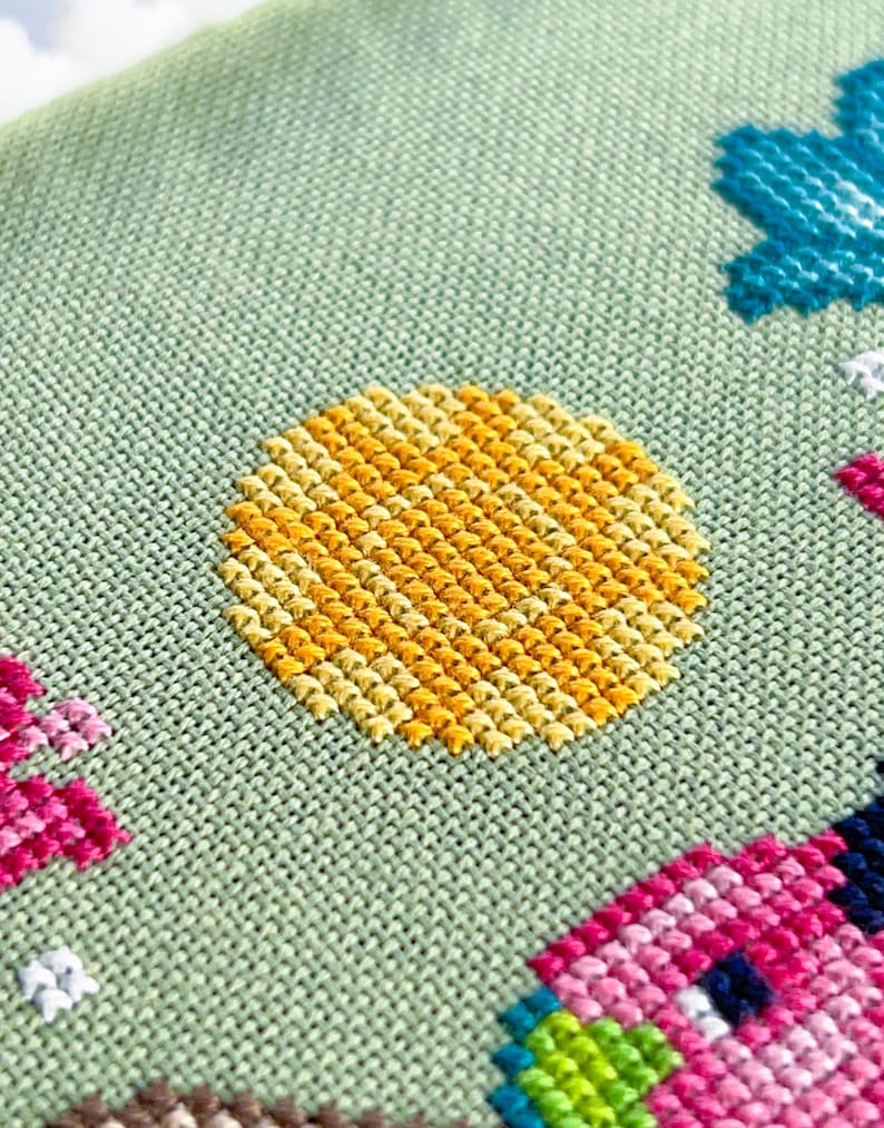 May include: Close-up of a cross-stitch design on a light green fabric. The design features a yellow sun, a blue flower, and a pink house. The stitches are neatly aligned, creating a textured and colorful pattern.