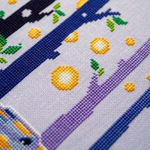 May include: Cross-stitch embroidery featuring a stylized design of branches and circular shapes in shades of blue, purple, yellow, and green on a light gray fabric. The design includes a partial image of a unicorn.