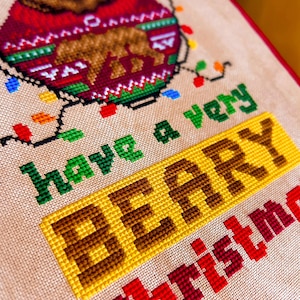 May include: Cross-stitch design featuring a bear wearing a red sweater with a bear graphic. The text "have a very BERRY Christmas" is stitched in green, yellow, and red. Colorful Christmas lights are also included in the design.