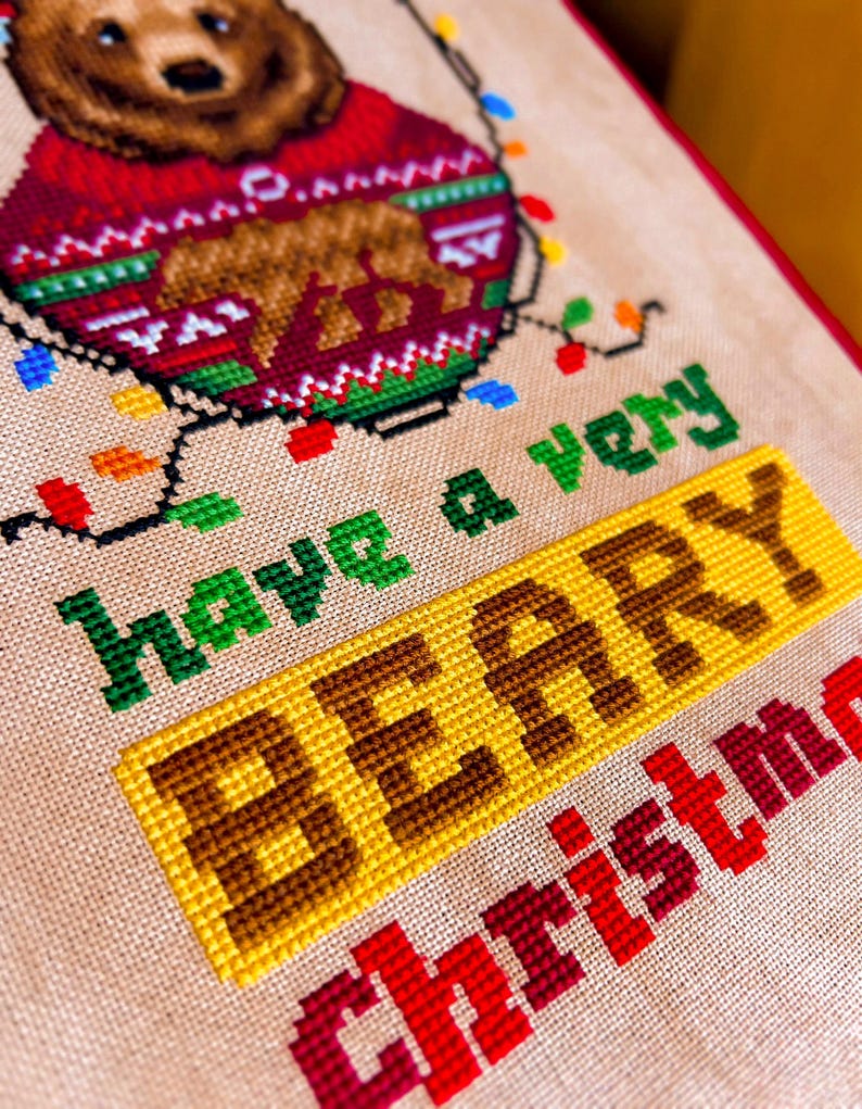 May include: Cross-stitch design featuring a bear wearing a red sweater with a bear graphic. The text "have a very BERRY Christmas" is stitched in green, yellow, and red. Colorful Christmas lights are also included in the design.