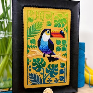 Może przedstawiać: Ramka z haftem krzyżykowym przedstawiająca tukan z jaskrawym pomarańczowym dziobem, niebieskimi i fioletowymi piórami oraz zielonymi tropikalnymi liśćmi na żółtym tle.