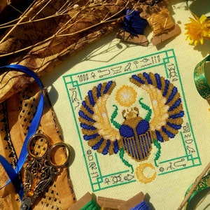 Top down view of Egyptian Scarab cross stitch pattern by NeedleLot Designs. Colors are blue, old gold, green, black and yellow. Piece consists only of DMC threads. Scarab is flanked by sun and moon, and is surrounded by hieroglyphs.