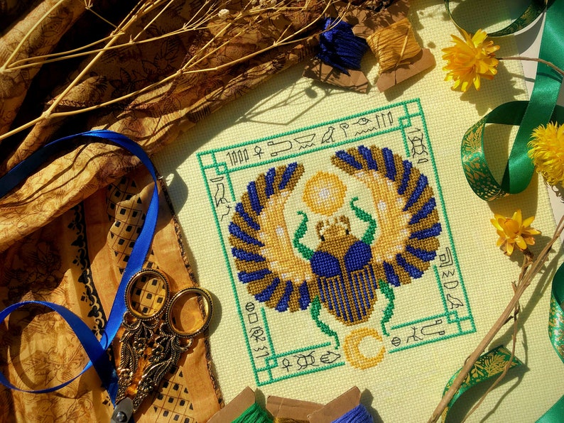 Top down view of Egyptian Scarab cross stitch pattern by NeedleLot Designs. Colors are blue, old gold, green, black and yellow. Piece consists only of DMC threads. Scarab is flanked by sun and moon, and is surrounded by hieroglyphs.