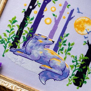 May include: Cross-stitch art of a wolf in a forest scene. The wolf is blue, purple, and yellow, lying down with a full yellow moon in the background. The trees are black, purple, and white with green leaves. The artwork is framed in a gold frame.