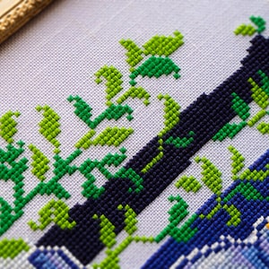May include: Close-up of a cross-stitch design featuring a navy blue and blue diagonal stripe pattern with green and light green leaves. The design is on a white fabric, with a gold-colored frame in the upper left corner.