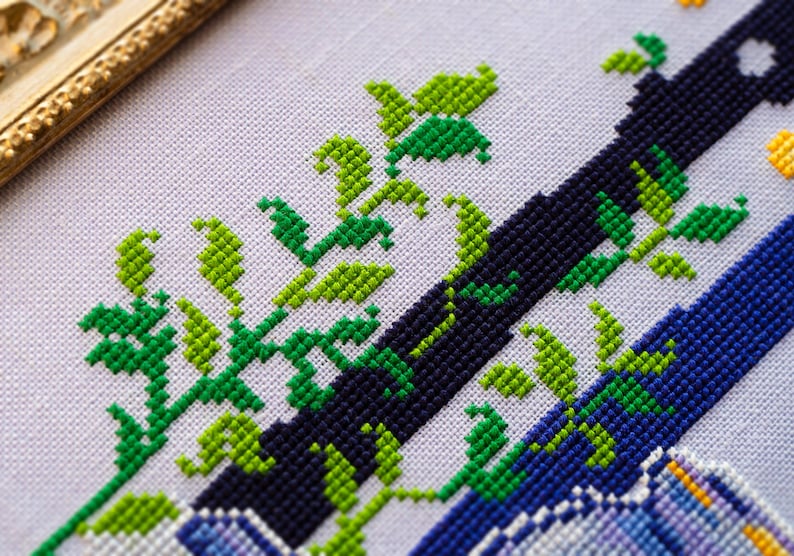 May include: Close-up of a cross-stitch design featuring a navy blue and blue diagonal stripe pattern with green and light green leaves. The design is on a white fabric, with a gold-colored frame in the upper left corner.
