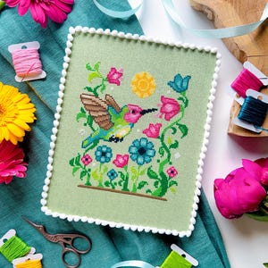 May include: A cross-stitch of a hummingbird with colorful wings, feeding from pink flowers, set against a light green background. The artwork is framed with a white pom-pom trim. Spools of thread and scissors are arranged around the artwork.