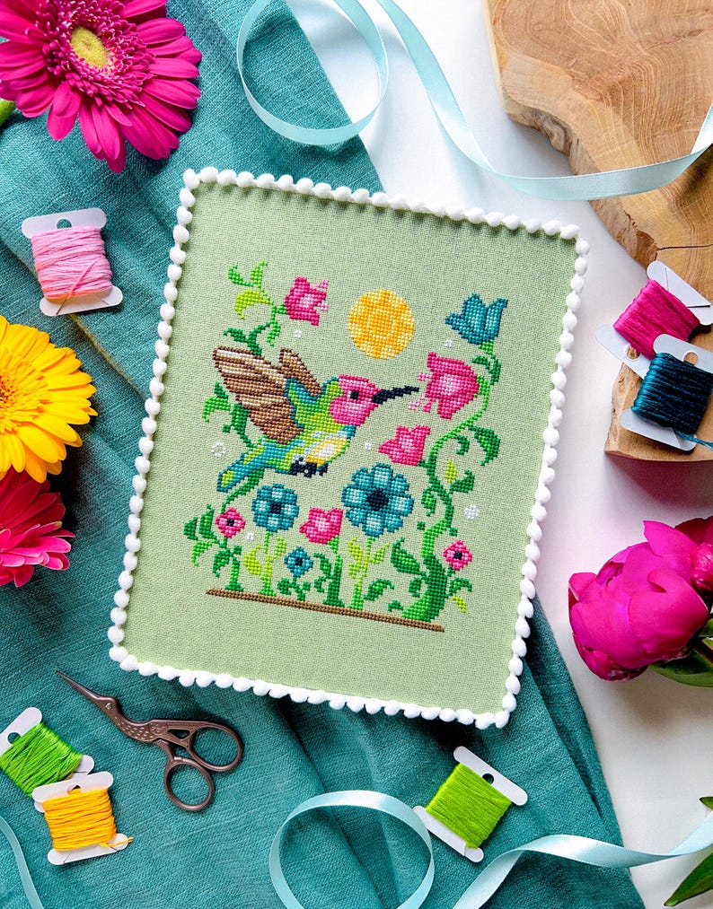 May include: A cross-stitch of a hummingbird with colorful wings, feeding from pink flowers, set against a light green background. The artwork is framed with a white pom-pom trim. Spools of thread and scissors are arranged around the artwork.