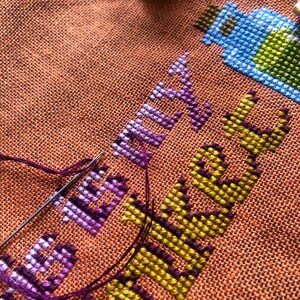 Closeup of the Trinket Bag cross stitch pattern being stitched. Stitches are neat and beautiful, colors really pop. Needle with thread is visible, and is placed on top of the work in progress.