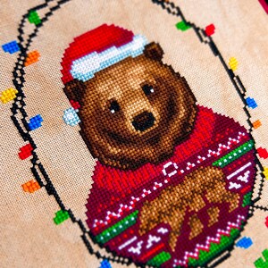 May include: Cross-stitch design of a brown bear wearing a red Christmas sweater and Santa hat. The bear is surrounded by a string of colorful Christmas lights, all stitched on a beige fabric background. The design has a festive holiday theme.
