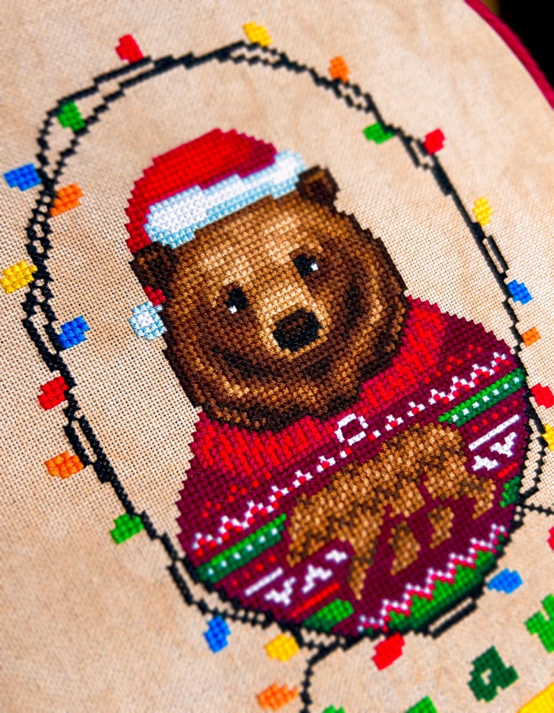 May include: Cross-stitch design of a brown bear wearing a red Christmas sweater and Santa hat. The bear is surrounded by a string of colorful Christmas lights, all stitched on a beige fabric background. The design has a festive holiday theme.