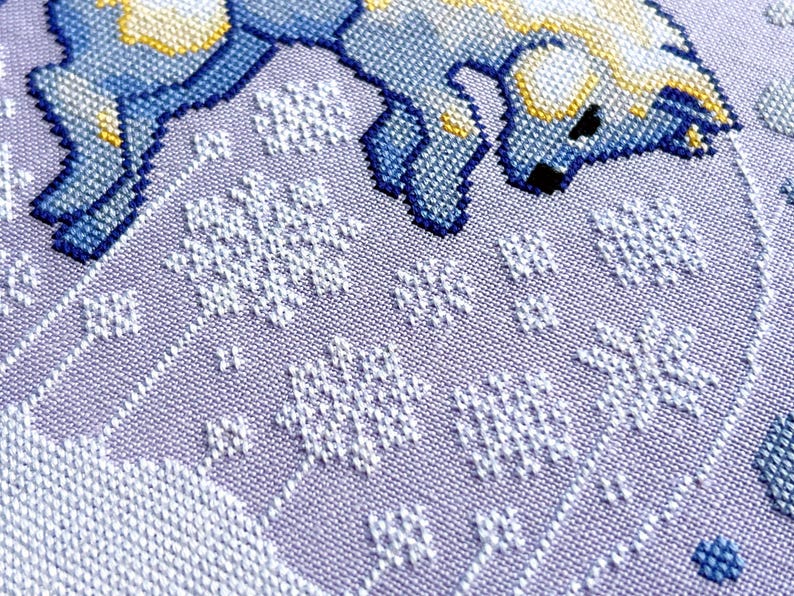 May include: A close-up of a cross-stitch pattern featuring a blue and yellow animal with white snowflakes on a purple background.
