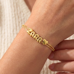 Peut inclure: Bracelet doré avec le nom "ZARA" en lettres majuscules et un charm en forme de cœur. Le bracelet est porté au poignet. Bijou de poignet.