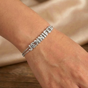 Puede incluir: Pulsera plateada con el nombre "Jennifer" escrito en letras mayúsculas. La pulsera está formada por una fina cadena y se lleva en la muñeca. El fondo es de color neutro.