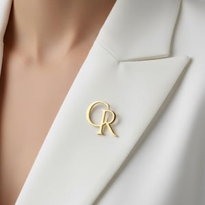 Op de afbeelding: Een goudkleurige speld in de vorm van de letters "CR" is bevestigd aan een witte blazer. De speld bevindt zich op de revers van de blazer. De blazer is gemaakt van een glad, mogelijk satijnachtig materiaal.