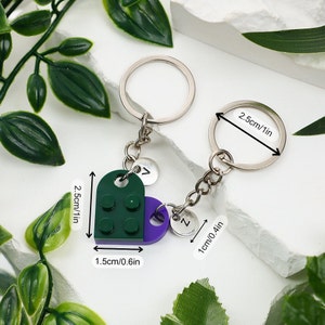 May include: A green and purple heart-shaped keychain with a silver chain and two silver charms. The charms are round and have the letters 'Z' and 'V' on them. The keychain measures 2.5 inches in length and 1.5 inches in width.
