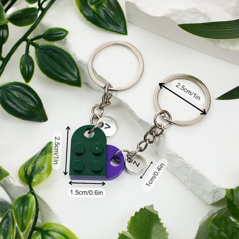 May include: A green and purple heart-shaped keychain with a silver chain and two silver charms. The charms are round and have the letters 'Z' and 'V' on them. The keychain measures 2.5 inches in length and 1.5 inches in width.