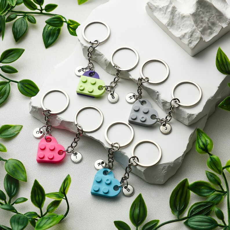 May include: Five silver keychains with heart-shaped charms made from Lego bricks. The charms are in different colors: green, gray, pink, and blue. Each keychain has a small silver charm with a letter on it.