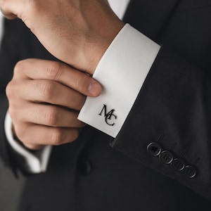 May include: A person adjusting a white dress shirt cuff with the black initials "MC". The cuff is part of a black suit jacket. The image focuses on formal attire and accessories, showcasing a classic style.