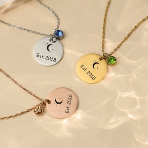 Peut inclure: Trois colliers en argent avec un pendentif en forme de lune et d'étoiles et un petit pendentif en pierre précieuse. Les colliers sont sur une surface blanche. Les breloques indiquent "Est. 2018".