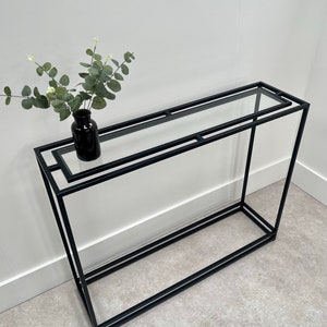 May include: A contemporary console table with a black metal frame and a clear glass surface. A small black vase contains eucalyptus sprigs. The table is set against a white wall. The table's dimensions are approximately 120cm in length.