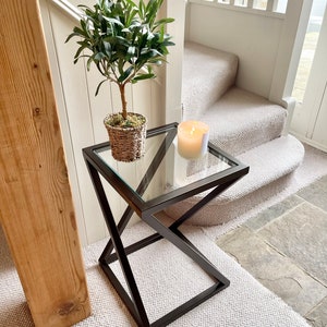 May include: A black metal side table with a glass top. The table has a geometric design and is sitting on a white carpet. A small potted plant and a candle are on the table.