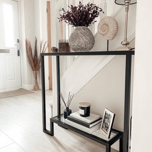 May include: A black metal console table with a glass shelf, displaying home decor. A textured vase with burgundy leaves, a seashell ornament, a black lamp, books, a candle, and a framed photo are visible.