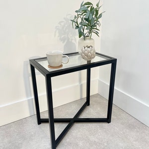 May include: A black metal side table with a glass top. The table has a criss-cross design on the legs and a small potted plant on the top.