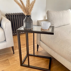 May include: A black metal side table with a glass top. The table has a simple, modern design and is ideal for a living room or bedroom. A white ceramic mug sits on the table, along with a decorative vase filled with dried pampas grass.