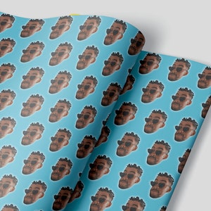 May include: Gift wrap featuring a repeating pattern of a person's face wearing heart-shaped sunglasses on a light blue background. The paper is folded over, revealing the pattern on both sides.