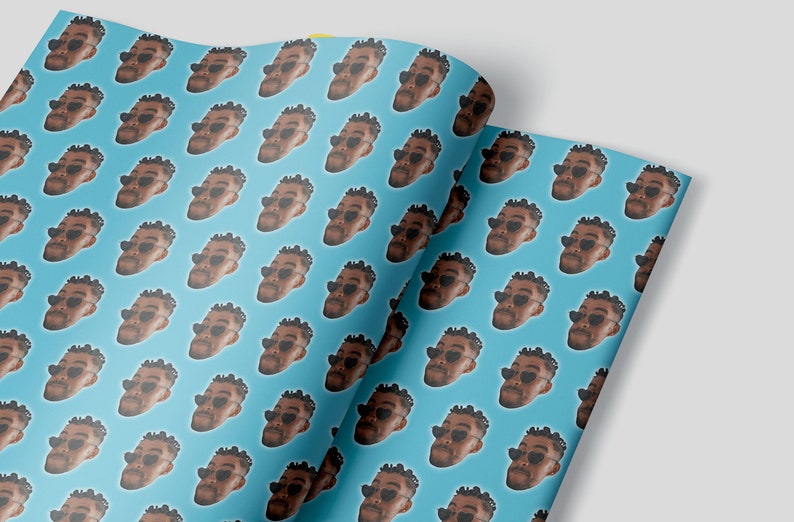 May include: Gift wrap featuring a repeating pattern of a person's face wearing heart-shaped sunglasses on a light blue background. The paper is folded over, revealing the pattern on both sides.