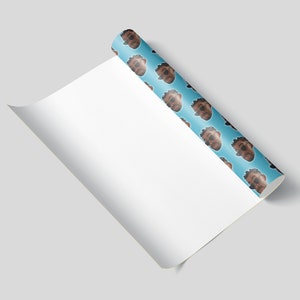 May include: A roll of wrapping paper with a repeating pattern of a person's face on a light blue background. The paper is partially unrolled, revealing a blank white surface. The image is set against a light gray background.