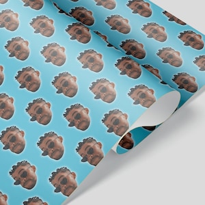 May include: A roll of wrapping paper with a repeating pattern of a person's face wearing sunglasses on a light blue background. The paper is partially unrolled, revealing the underside.