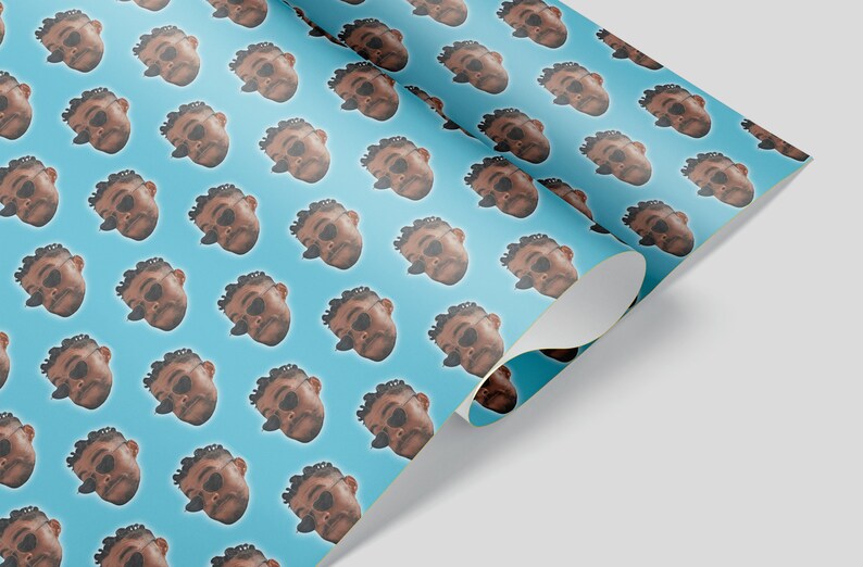 May include: A roll of wrapping paper with a repeating pattern of a person's face wearing sunglasses on a light blue background. The paper is partially unrolled, revealing the underside.